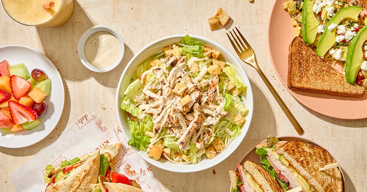 Spread of Rachel's Kitchen dishes including a grilled chicken salad, sandwiches, fresh fruit, avocado toast, and a smoothie
